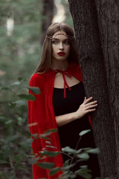 Beautiful mystic girl in red cloak and elegant wreath in forest