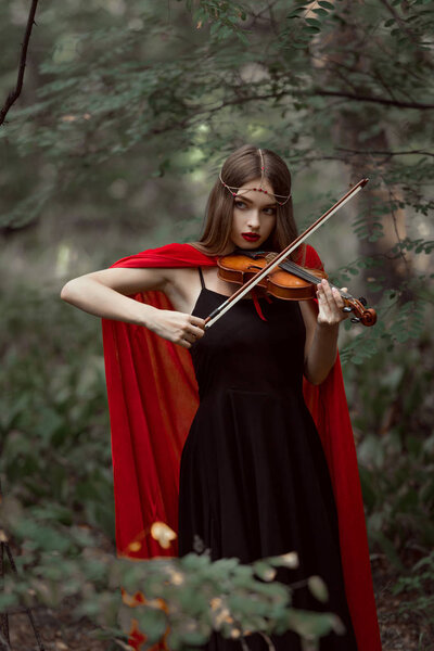 beautiful mystic girl in red cloak playing on violin in dark woods