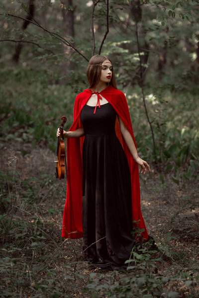 Elegant mystic girl in black dress and red cloak holding violin in dark woods