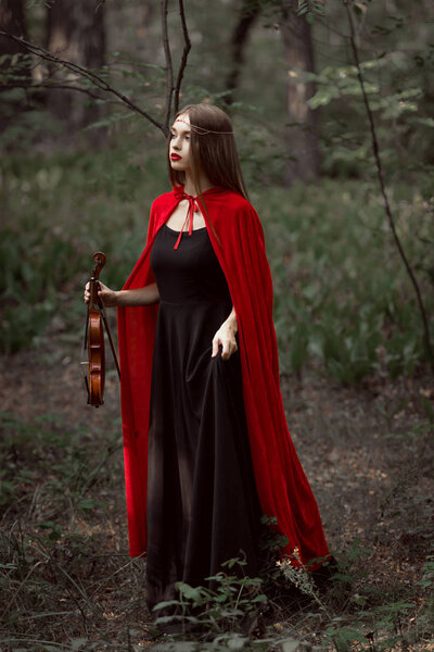 Attractive mystic woman in red cloak holding violin in dark forest
