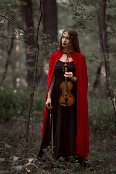 elegant beautiful woman in black dress and red cloak holding violin in forest