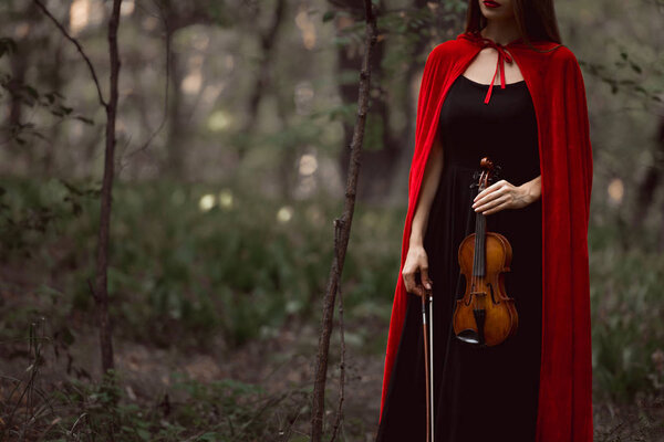 Cropped view of elegant woman in red cloak holding violin in dark woods