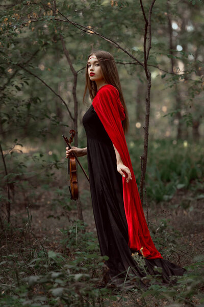 attractive mystic woman in black dress and red cloak holding violin in forest
