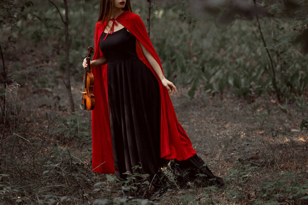 cropped view of elegant mystic woman in black dress and red cloak holding violin in forest