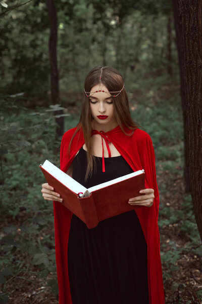 Beautiful mystic girl in red cloak and wreath reading magic book in woods