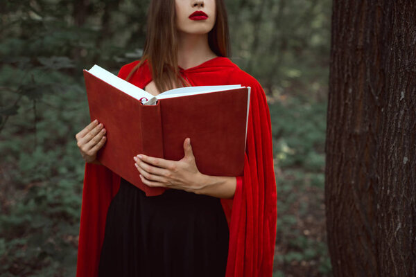 Cropped view of girl in red cloak holding magic book in forest