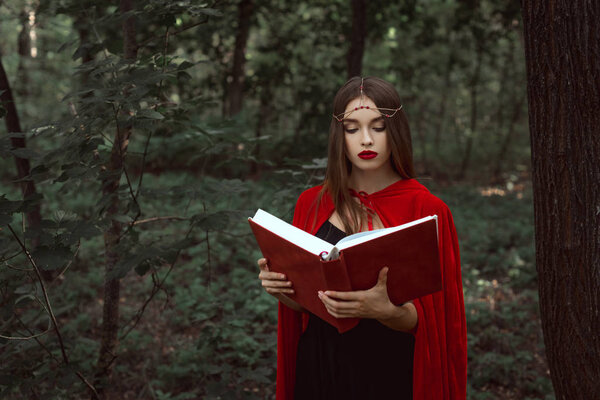 attractive mystic woman in red cloak and wreath reading magic book in forest