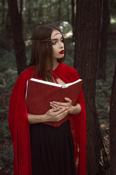 красивая мистическая девушка в красном плаще и венке держит волшебную книгу в лесу
