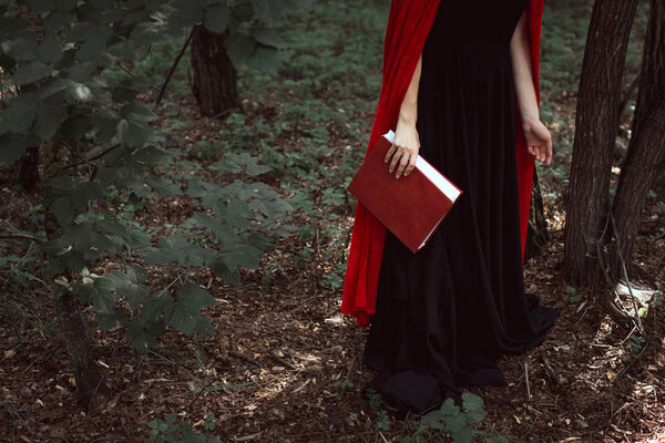Cropped view of elegant girl in red cloak with magic book in forest