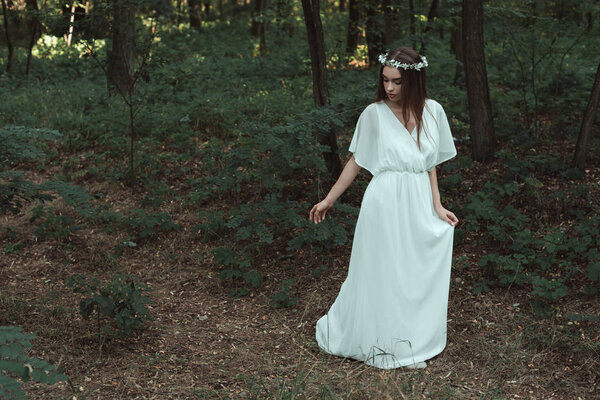 Elegant girl in white dress and floral wreath walking in woods
