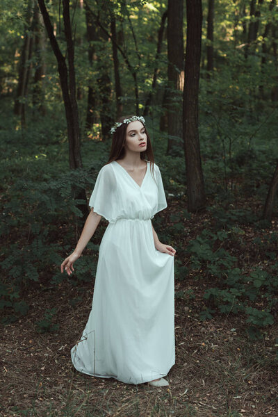 Pretty girl in elegant white dress and floral wreath walking in woods