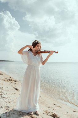 beautiful elegant girl in white dress and floral wreath playing violin on sand beach clipart