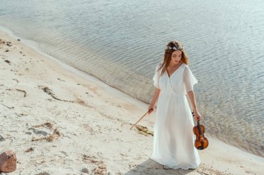 attractive girl in white dress holding violin on seashore