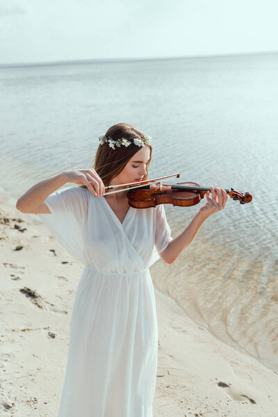 beautiful woman in elegant white dress and floral wreath playing violin on seashore