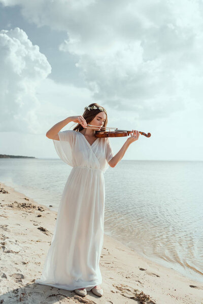 beautiful elegant girl in white dress and floral wreath playing violin on sand beach