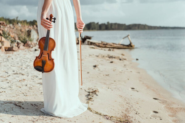 cropped view of elegant girl in white dress holding violin on sand beach
