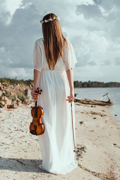 back view of girl in elegant white dress holding violin on seashore