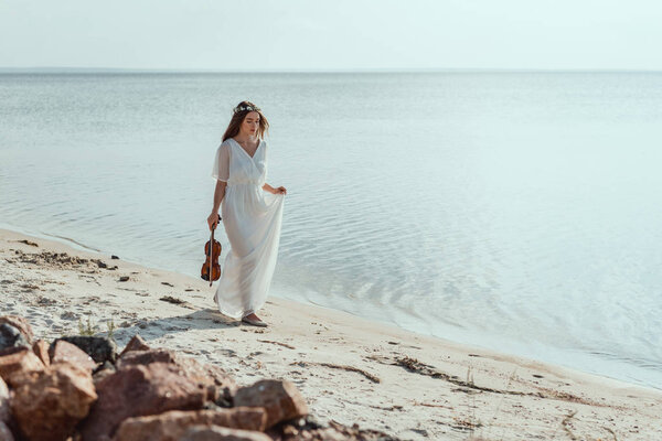 attractive young woman in elegant dress with violin walking on beach near sea
