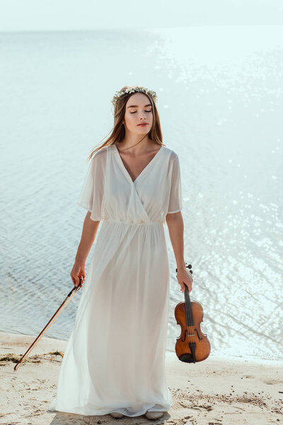 attractive woman with closed eyes posing in elegant dress and holding violin near sea