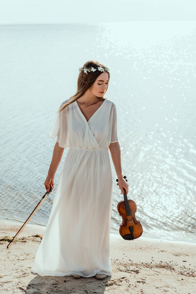 beautiful elegant woman in dress and floral wreath holding violin on beach near sea