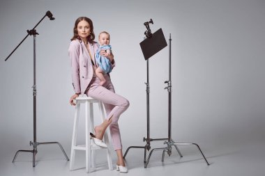 beautiful stylish mother holding adorable infant daughter and looking at camera while sitting on stool in recording studio clipart