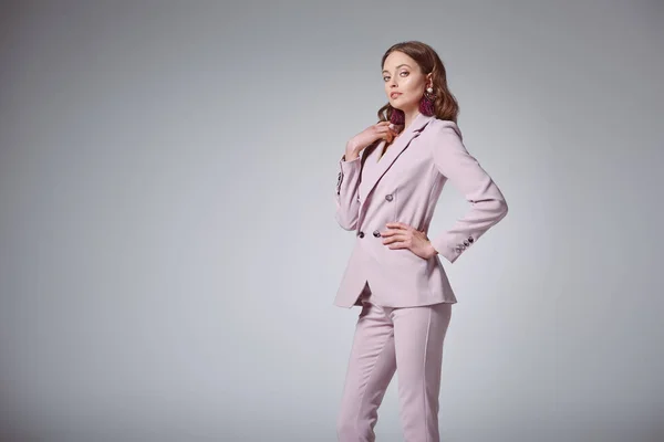 beautiful woman in stylish pink suit posing with hand on waist and looking at camera isolated on grey