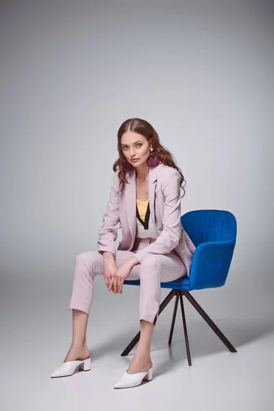 fashionable woman in stylish pink suit sitting on chair and looking at camera on grey