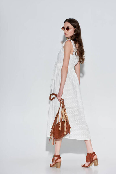 side view of beautiful woman in stylish white dress and sunglasses holding handbag on grey
