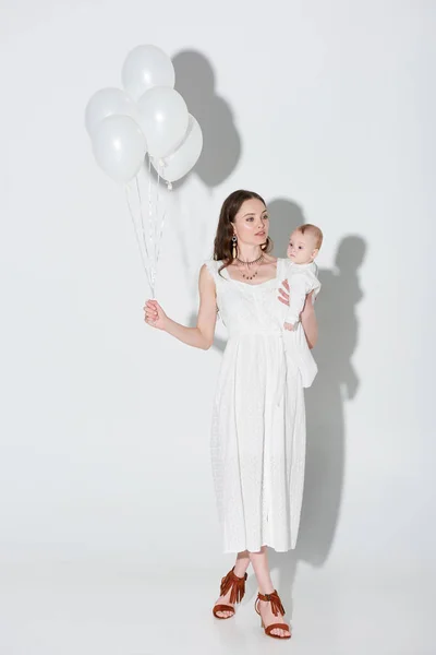 スタイリッシュな白いドレス女の愛らしい赤ちゃんを運ぶと灰色の風船の束を保持で美しい女性 — ストック写真