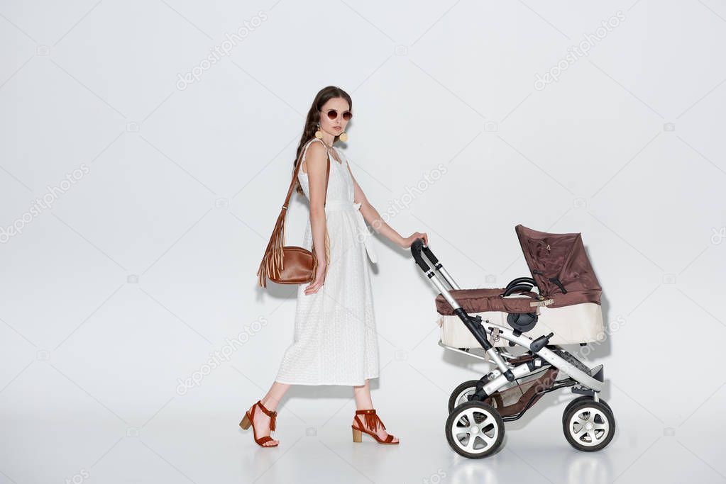 fashionable woman in white dress and sunglasses walking with baby carriage and looking at camera on grey 