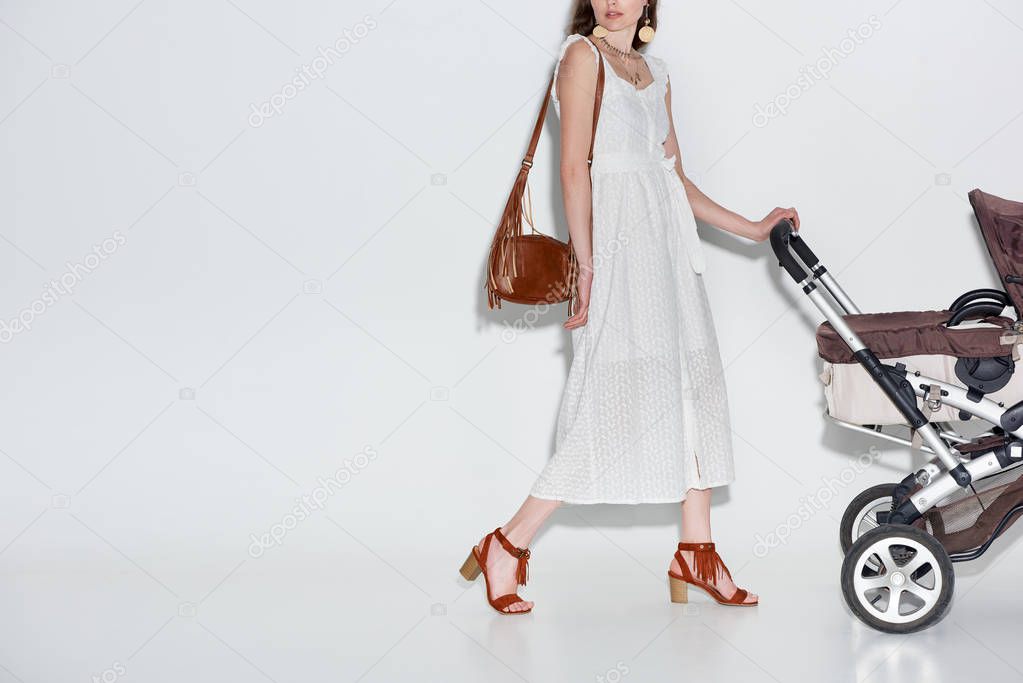 cropped shot of young woman in fashionable white dress walking with baby carriage on grey