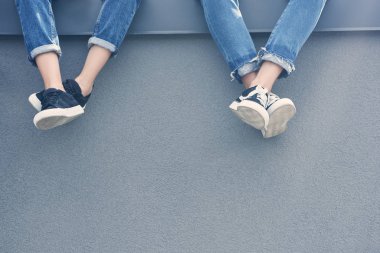 partial view of couple of legs in jeans and sneakers sitting on grey wall clipart