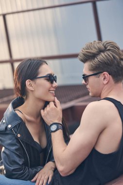 interracial happy couple in sunglasses flirting and looking at each other, man touching chin of girlfriend clipart