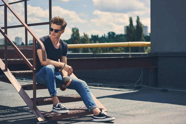 stylish man in sunglasses holding bottle of whiskey while sitting on stairs on roof