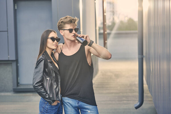 мультикультурная пара в модных солнцезащитных очках, обнимающаяся на крыше с подсветкой, пьющий пиво мужчина
