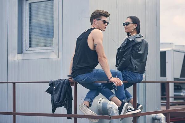 beautiful multicultural couple listening music with earphones while sitting on roof
