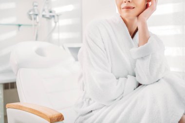 partial view of woman in white bathrobe sitting on massage table in spa salon clipart