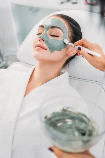 partial view of cosmetologist applying clay mask on female face in spa salon