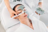 Teilansicht des Kosmetologen, der im Wellness-Salon Tonmaske mit Pinsel auf das weibliche Gesicht aufträgt