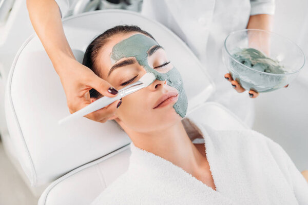 partial view of cosmetologist applying clay mask with brush on female face in spa salon