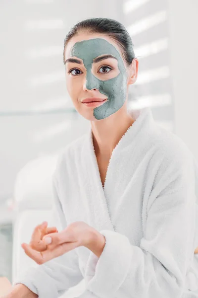 portrait of beautiful woman with clay mask on face in white bathrobe in spa salon