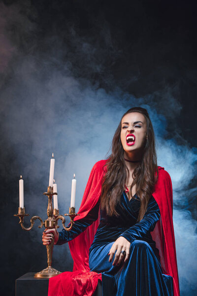 woman in vampire costume holding vintage candelabrum on darkness with smoke 