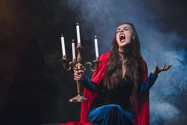 ビンテージの燭台を押し 煙で暗い背景に吸血鬼の牙を示す赤いマントの女性 — ストック写真