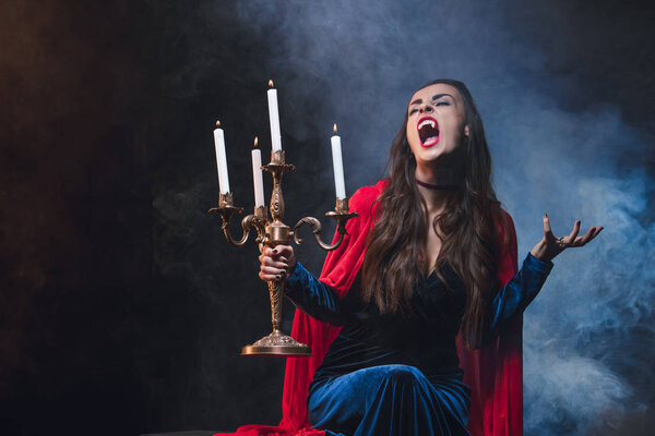 woman in red cloak holding vintage candelabrum and showing vampire fangs on dark background with smoke 