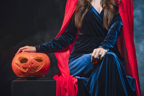 cropped view of woman in red cloak with jack o lantern on darkness with smoke 