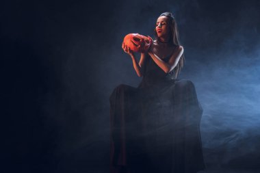 beautiful woman in vampire costume holding jack o lantern on dark background with smoke  clipart