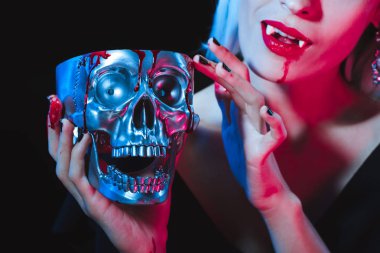 cropped view of vampire holding metal skull with blood isolated on black clipart