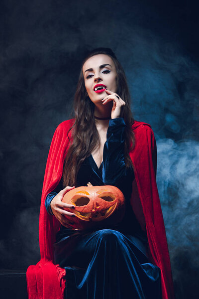 attractive woman in vampire costume holding jack o lantern on darkness with smoke 