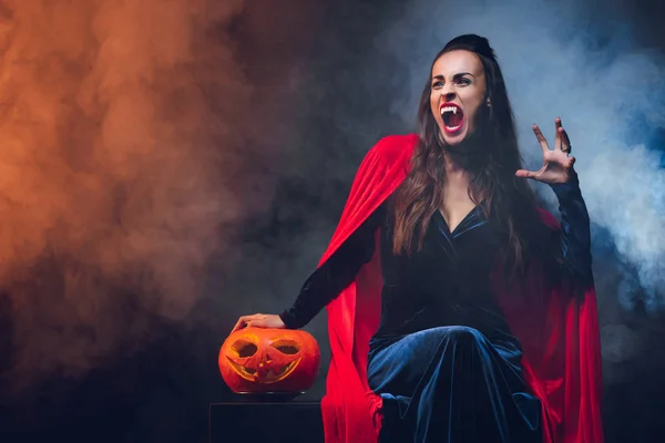 mystic woman in vampire costume holding jack o lantern on darkness with smoke