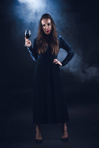 grim vampire holding wineglass with blood on dark background with smoke 